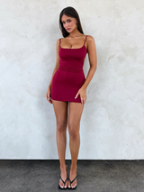 A woman poses against a textured white wall wearing the TIGERMIST MINERVA MINI DRESS in red and black heeled sandals. Her hair is straight, and she stands with one hand on her hip, looking at the camera.