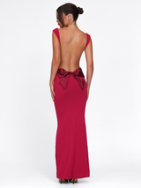 A woman stands facing away in a TIGERMIST ZITA DRESS - RED : SALSA : DEEP RED, a sleeveless, floor-length bodycon dress featuring an open back with a large bow, her hair styled in a neat bun against a plain white background.