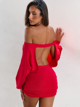 A woman stands against a white wall wearing the TIGERMIST JANINE MINI DRESS in red, featuring an off-shoulder neckline, open back, and ruched skirt, as she looks down over her shoulder.
