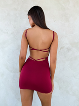 A woman with long brown hair and large hoop earrings poses against a light textured wall, wearing the TIGERMIST MINERVA MINI DRESS in red—a sleeveless, backless style with thin adjustable straps.